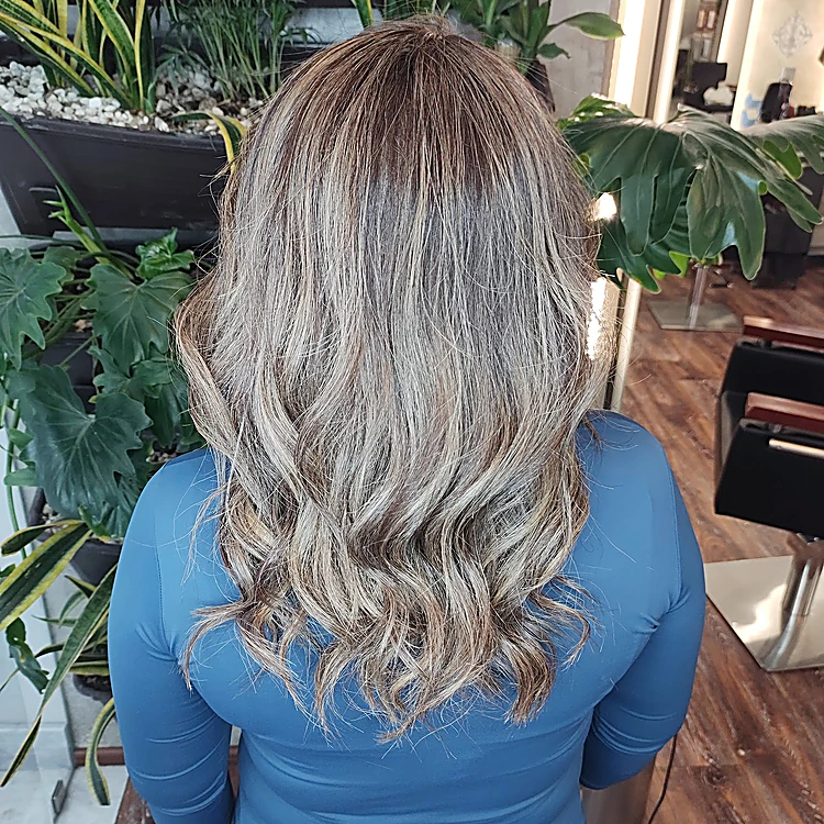 Clienta de espaldas con balayage ceniza en ondas, frente a una jardinera con monstera y sansevieria