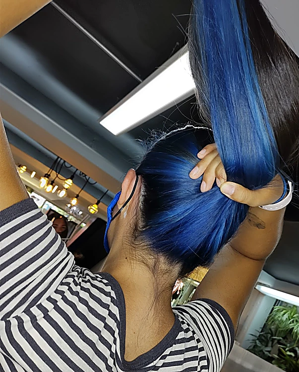Estilista sostiene en alto un mechón teñido de azul eléctrico sobre cabello oscuro