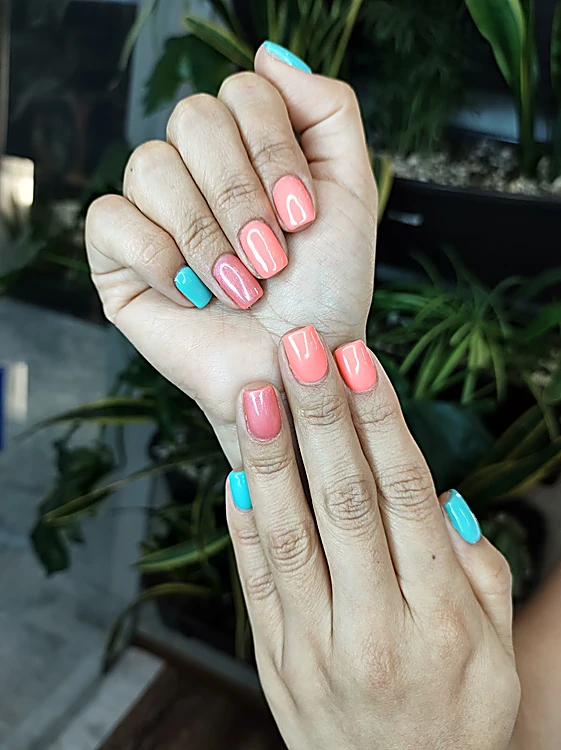 Manos con uñas coral y una turquesa sobre fondo de hojas verdes
