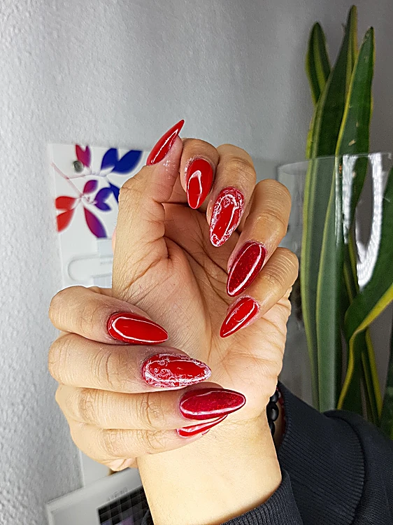 Mano con uñas largas en punta, rojas con acabado cromado, junto a una planta